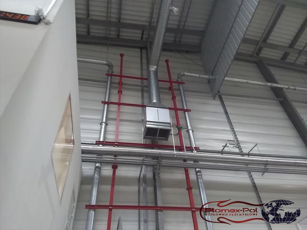 Nagrzewnica powietrza zamontowana na konstrukcji nośnej w hali przemysłowej z widocznymi rurami instalacyjnymi.