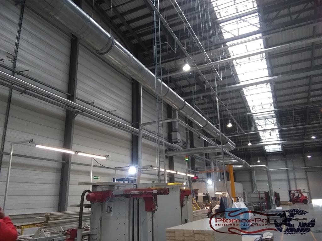 Szeroki widok instalacji rur doprowadzających ciepłe powietrze do maszyn wewnątrz hali produkcyjnej.