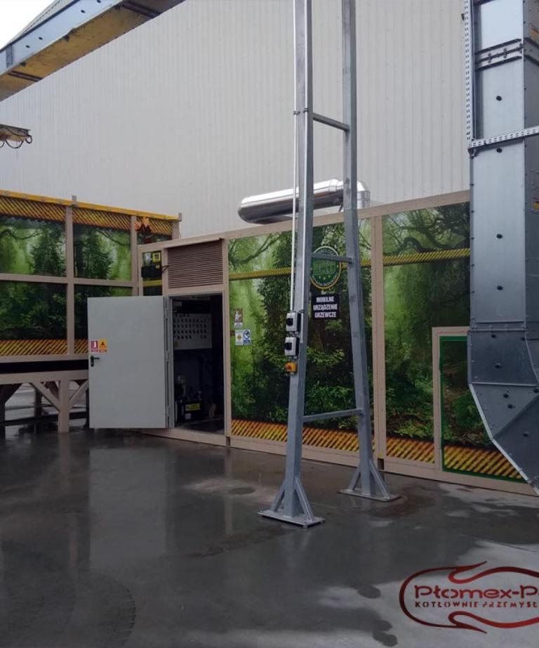Mobilna, kontenerowa kotłownia przemysłowa z dekoracyjną grafiką przedstawiającą las, zamontowana przy budynku hali fabrycznej. Widoczny jest system podajników i metalowe przewody kominowe, a w prawym dolnym rogu logo Plomex-Pol.